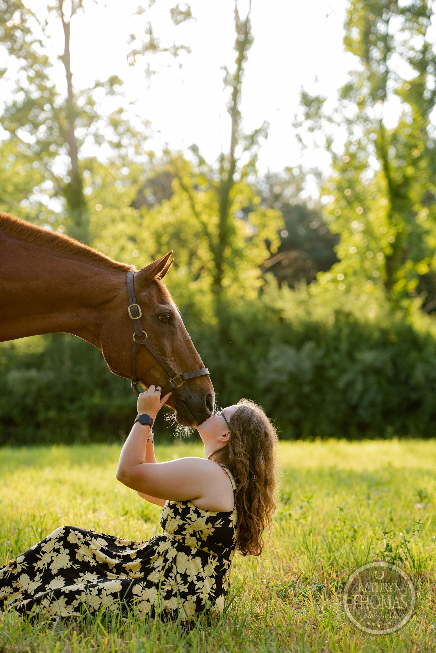 Someone in a floral dress lies in grass reaching up to pet a chestnut horse's nose in golden sunlight.
