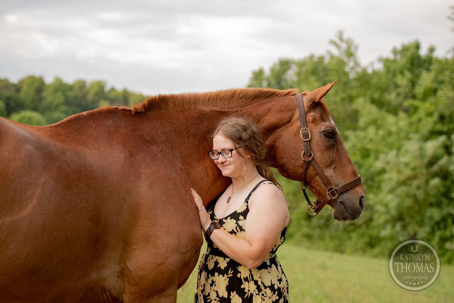 A chestnut horse stands calmly beside its handler in a floral dress on a summer day.