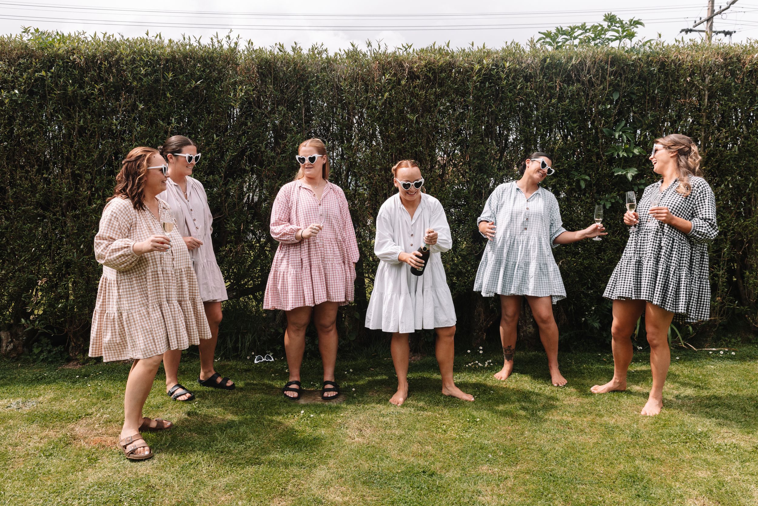 Bridesmaids in pastel dresses laughing and dancing together on a lawn with a hedge backdrop.
