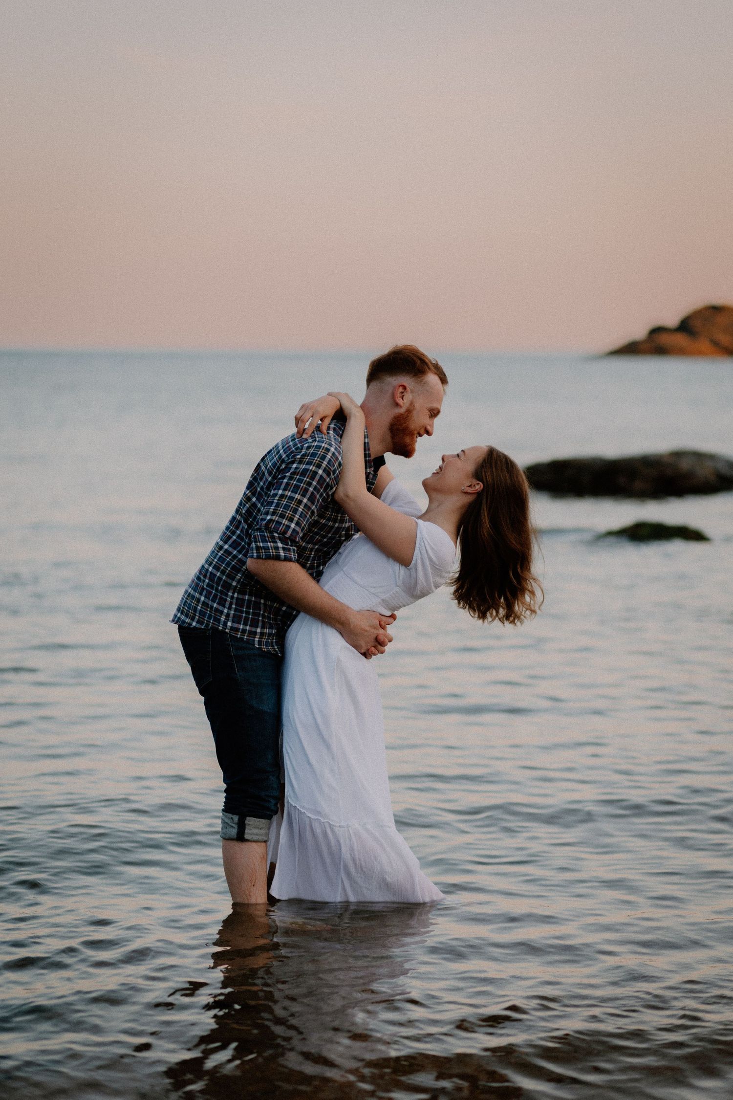 A romantic couple embraces in the ocean at sunset with waves lapping at their legs.