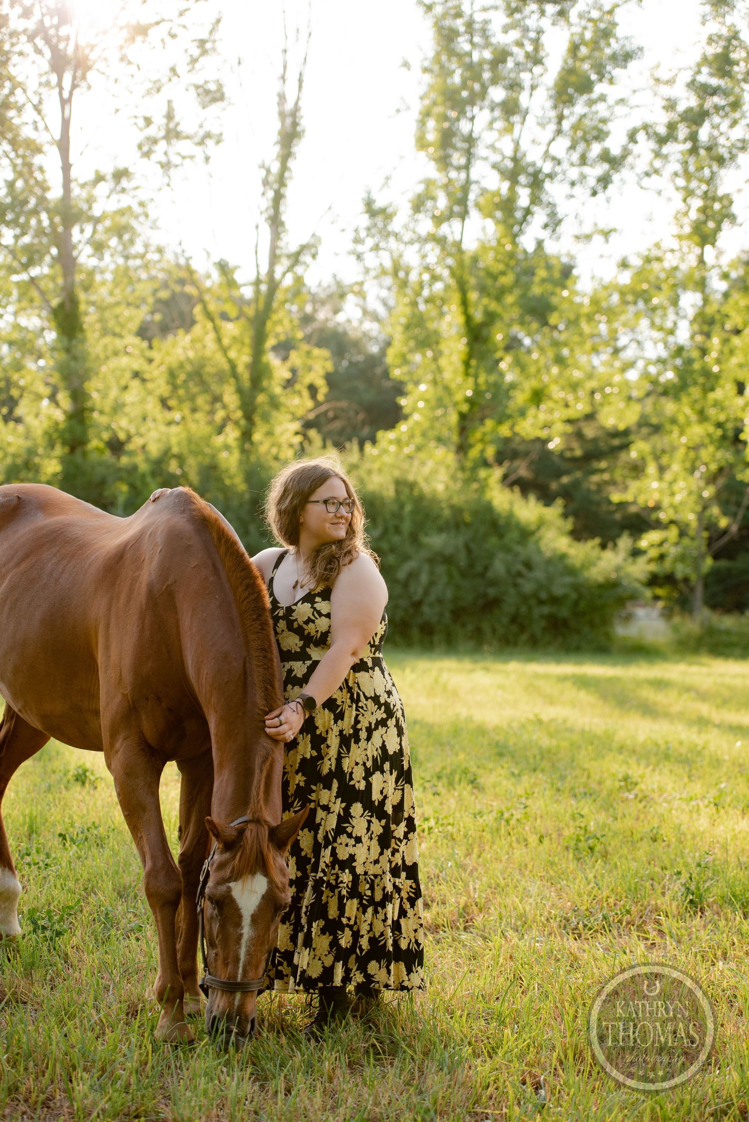 A chestnut horse and someone in a floral dress pose together in a sunlit field with tall trees.