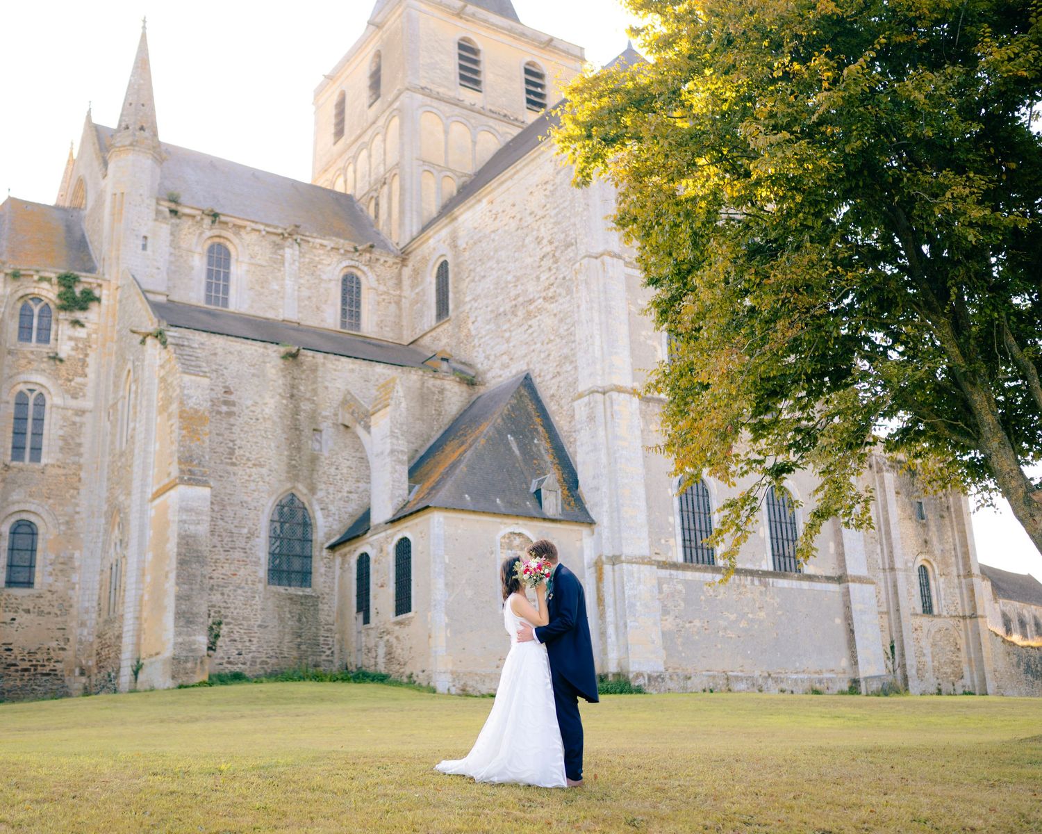 A wedding couple poses near a historic stone church with autumn trees and Gothic architecture in the background.