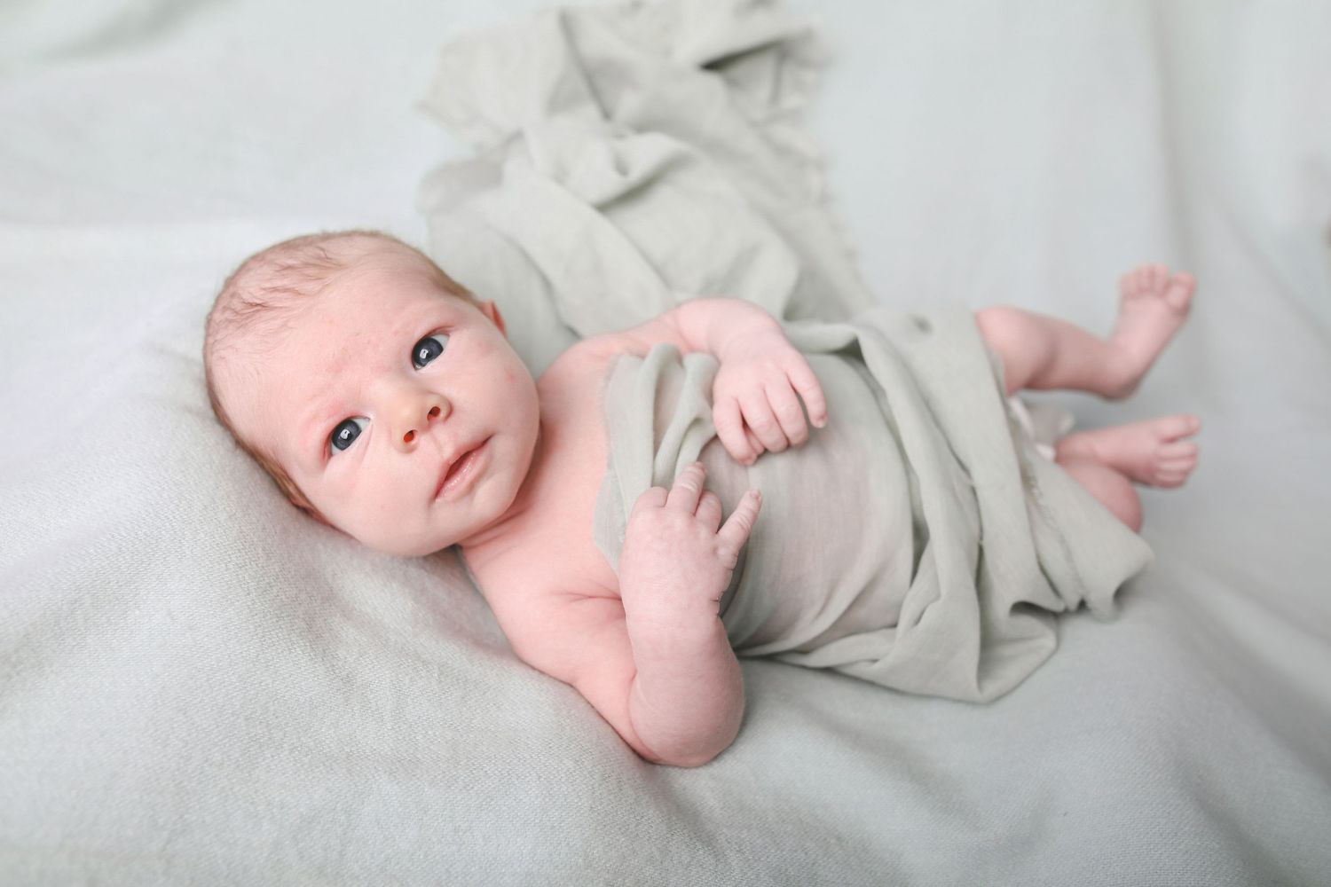 Infant wrapped in light gray fabric lying on white background with bright expression.