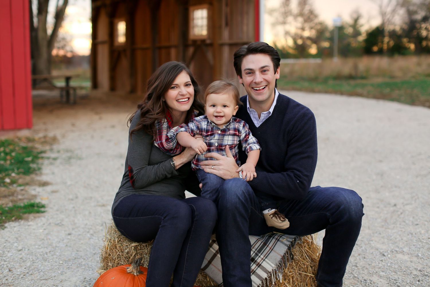 Autumn family portrait on hay bale with red barn backdrop during sunset.