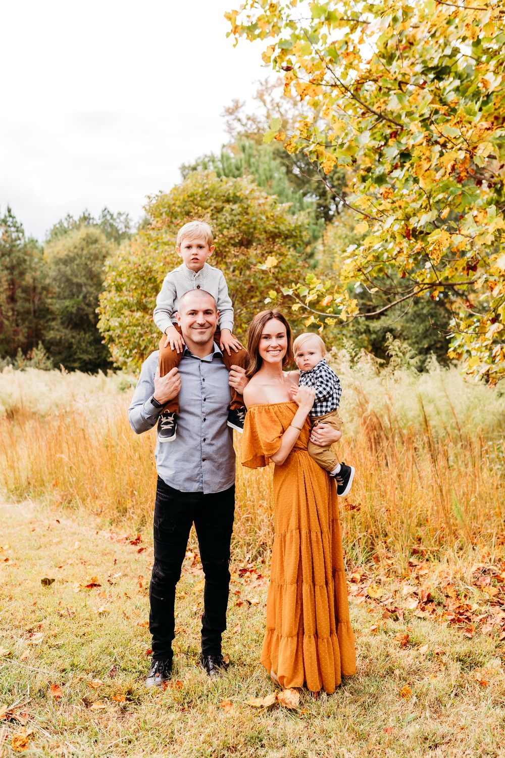 A family in coordinated autumn outfits poses together in a golden field with fall foliage in the background.