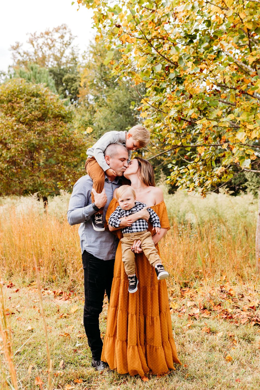 A family shares a tender autumn moment together in a field with golden grass and yellow-leaved trees.