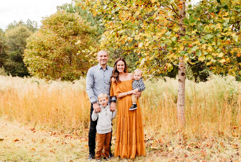 A couple stands together in a golden autumn field with tall grass and yellow foliage in the background.