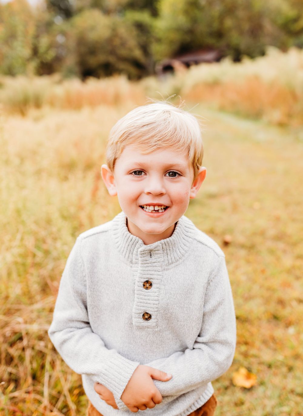 Child wearing a cozy gray knit sweater smiles warmly in an autumn outdoor setting with golden foliage in background.