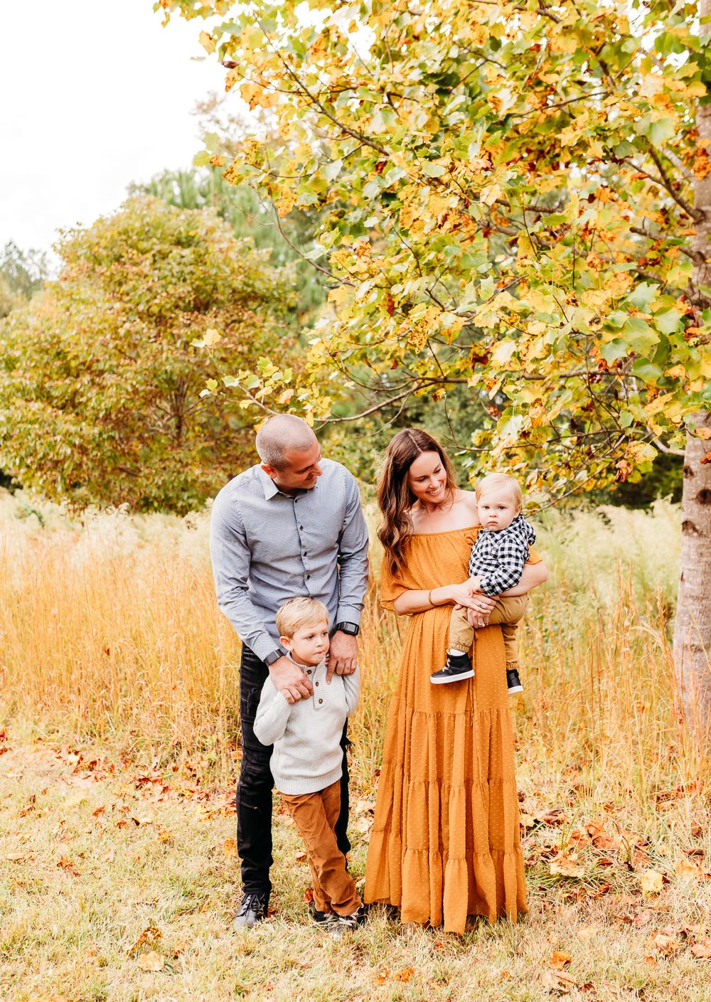 A family poses together in a golden autumn field with fall foliage and tall grass in the background.