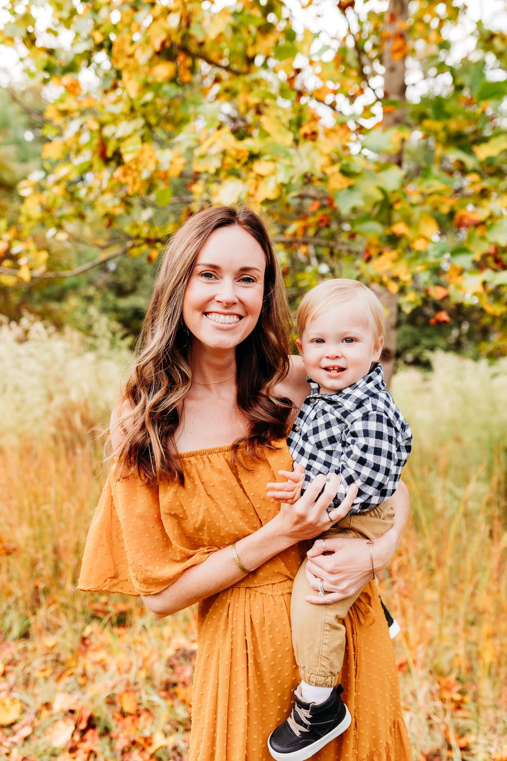 A person in a mustard yellow dress holds a small child wearing black and white against an autumn backdrop of golden leaves.