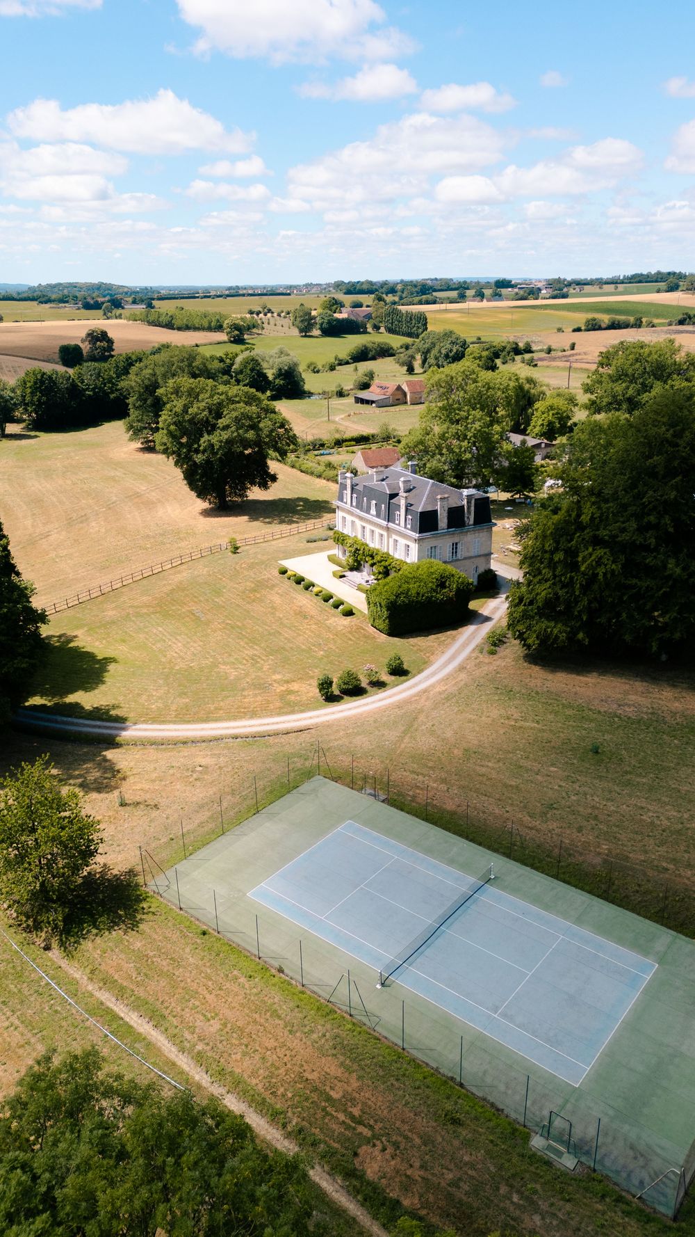 Aerial view of a private tennis court next to a white mansion surrounded by rolling countryside and mature trees.
