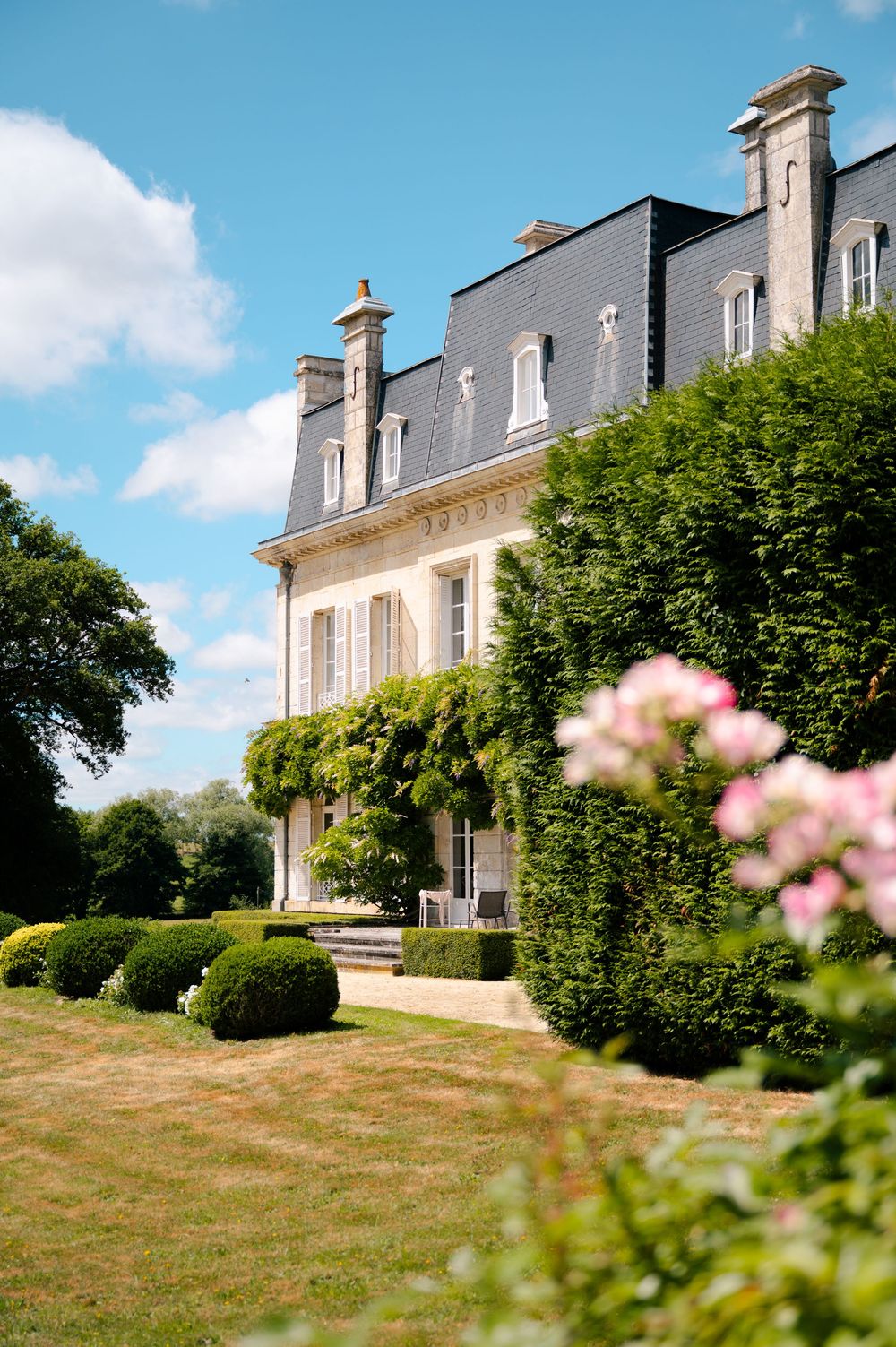 A panoramic view of a stately French chateau with mansard roof and manicured lawn under a bright blue summer sky.