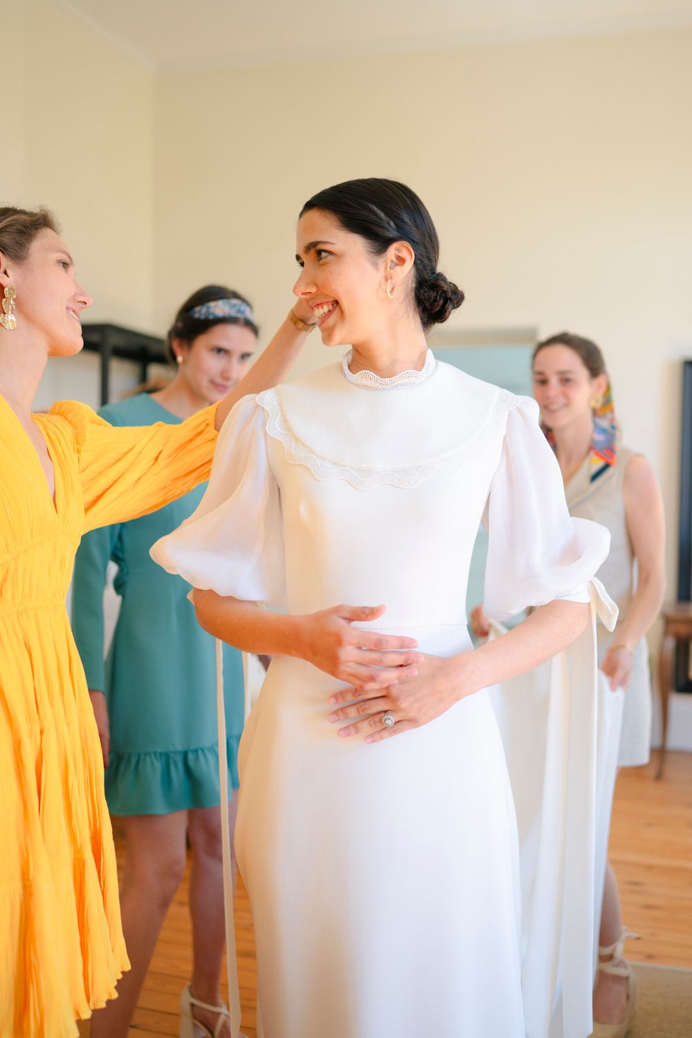 A bride in a sleek white wedding gown stands with attendants in yellow and teal dresses during pre-ceremony preparations.