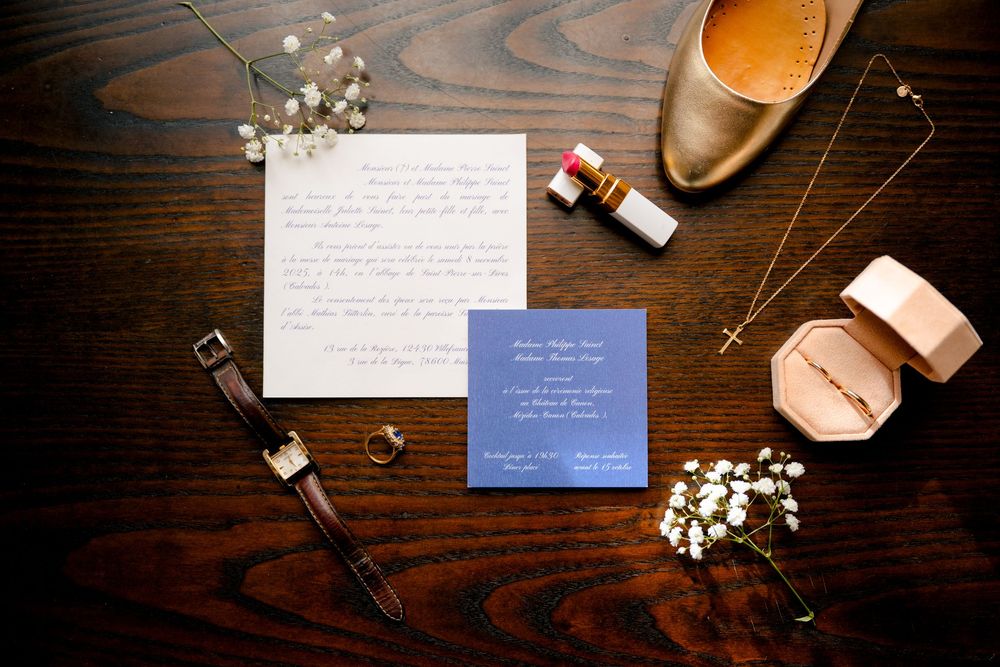 Wedding details including invitation, watch, shoes, flowers and makeup arranged on dark wooden surface.