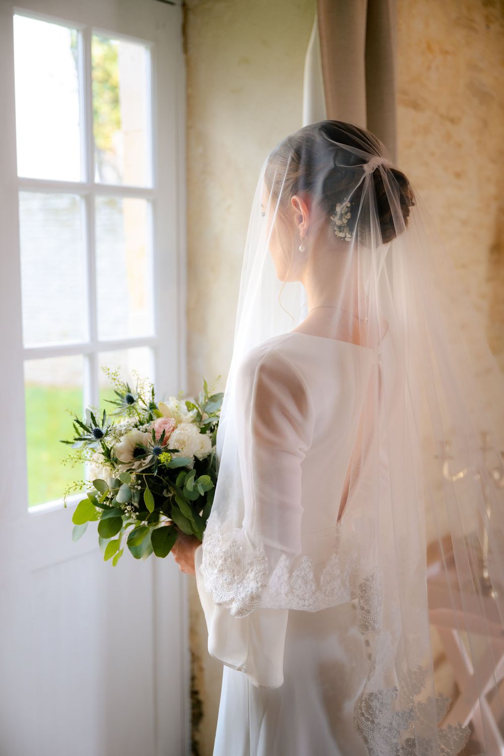 Ethereal bridal portrait series showing a veiled bride in white near sunlit windows with delicate floral bouquet.