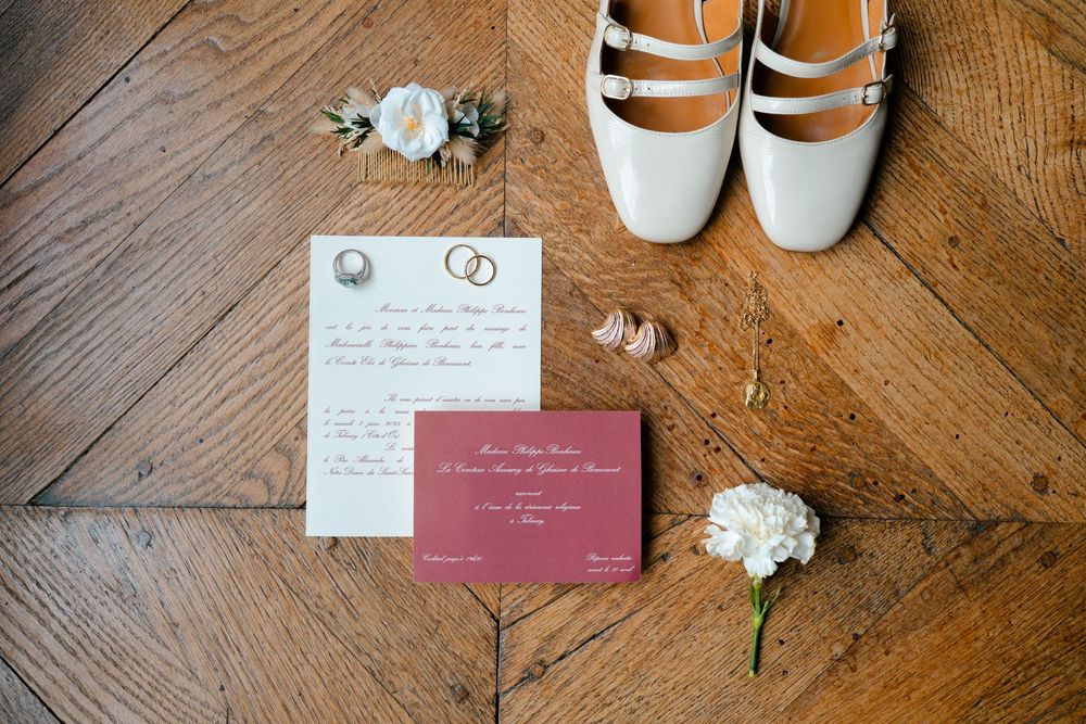 Wedding accessories including white shoes, floral details, and invitation cards arranged on a wooden floor.
