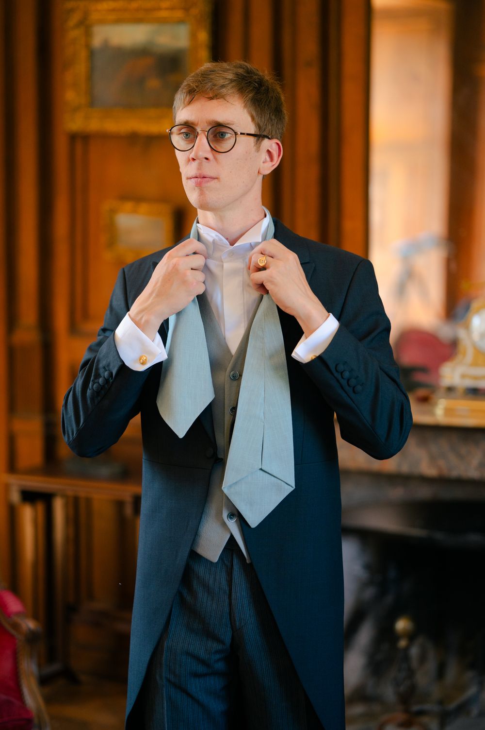 A person in formal attire adjusts their light blue tie while wearing a navy suit jacket and waistcoat in an elegant room.