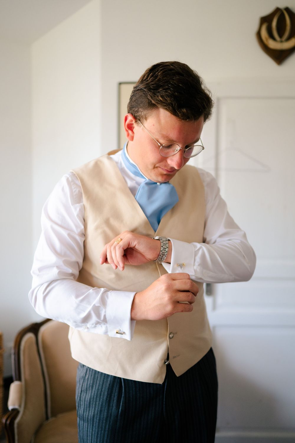 A person in formal attire adjusts their cuff links while wearing a beige vest, white shirt and light blue tie.