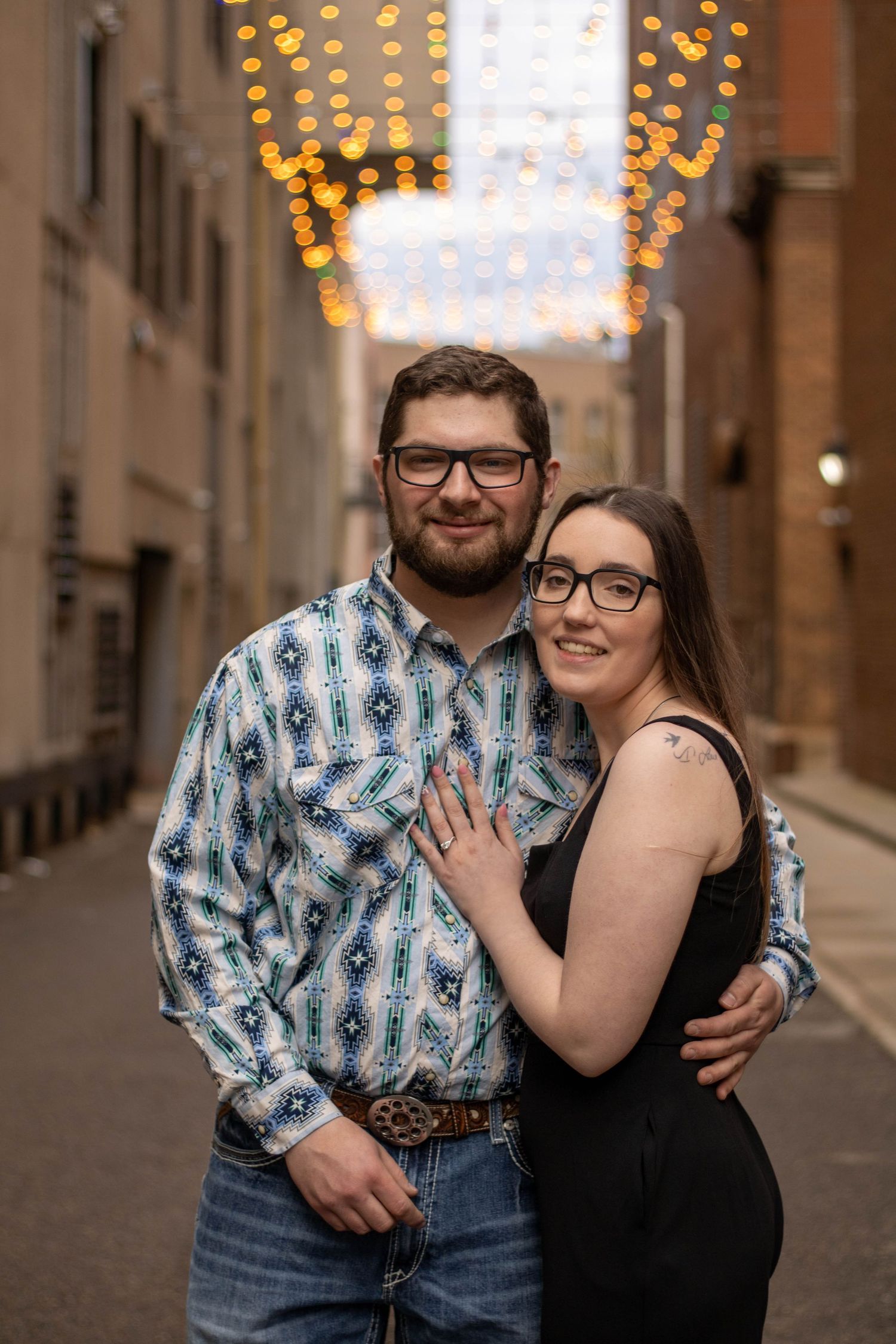 A couple in coordinating outfits share a romantic moment on a cobblestone street with string lights glowing overhead.