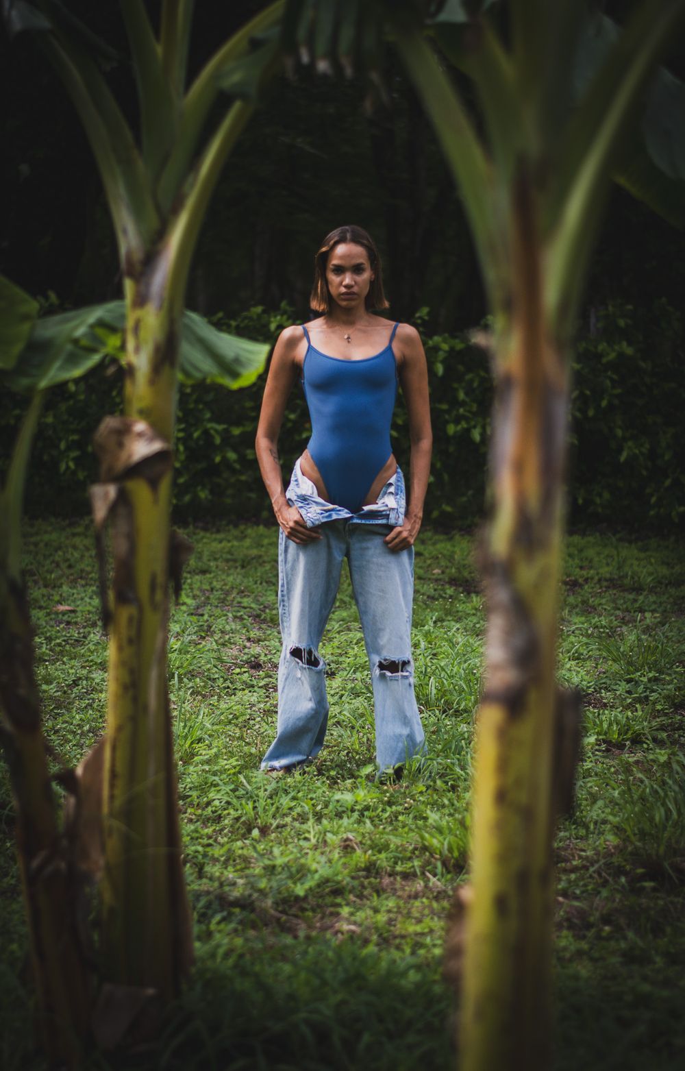 Blue bodysuit and distressed jeans outfit framed by tropical banana plants.