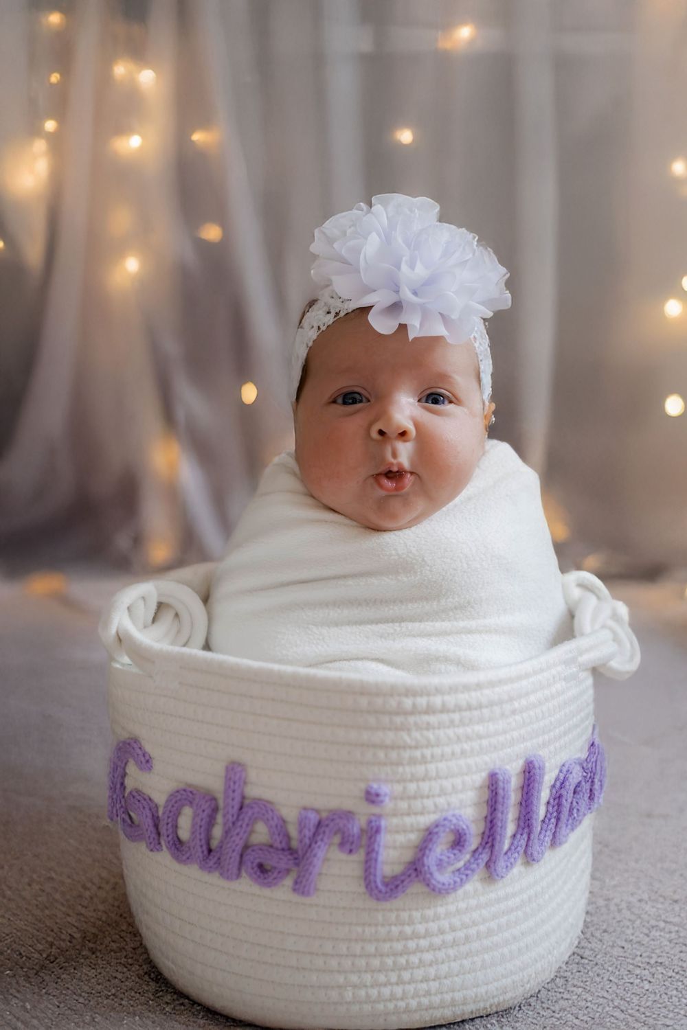 Newborn baby girl Gabriella sitting in personalized white rope basket with white flower headband and bokeh lights.