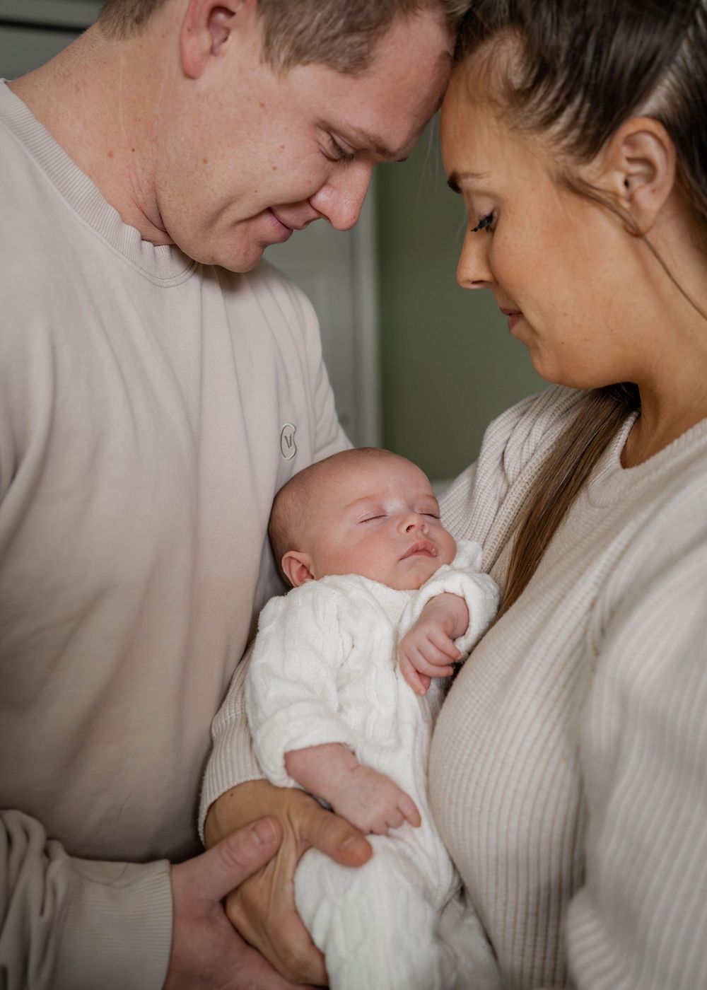 New parents in matching cream outfits tenderly gazing at sleeping newborn baby dressed in white.
