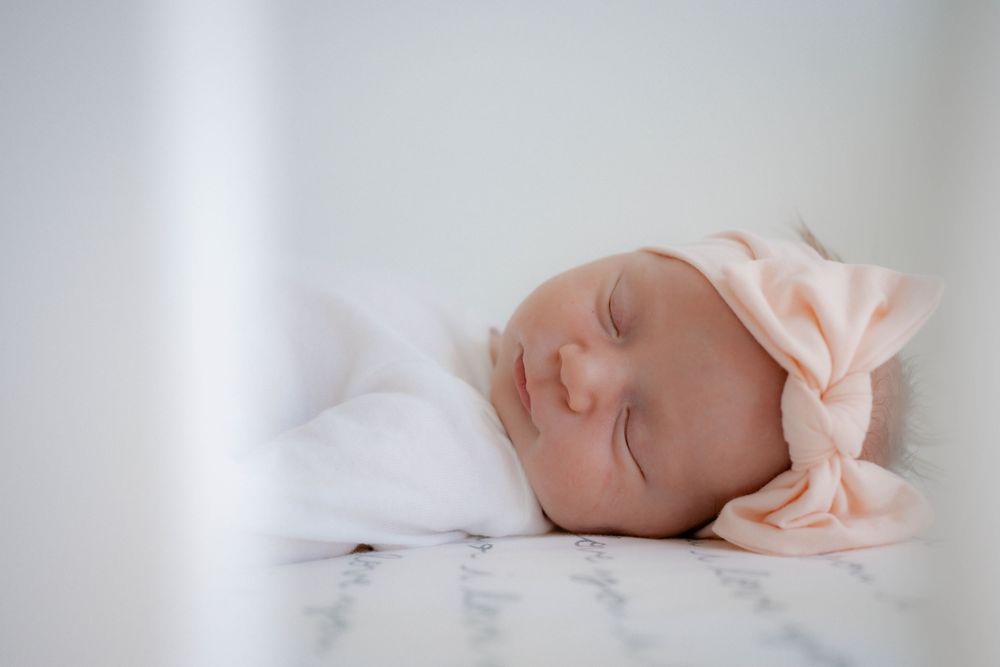 Peaceful newborn baby girl sleeping swaddled in white with pink bow headband in white crib.