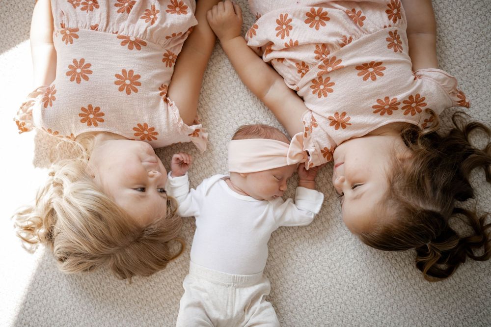 Two young siblings in floral dresses lying beside newborn baby girl with pink headband on white blanket.
