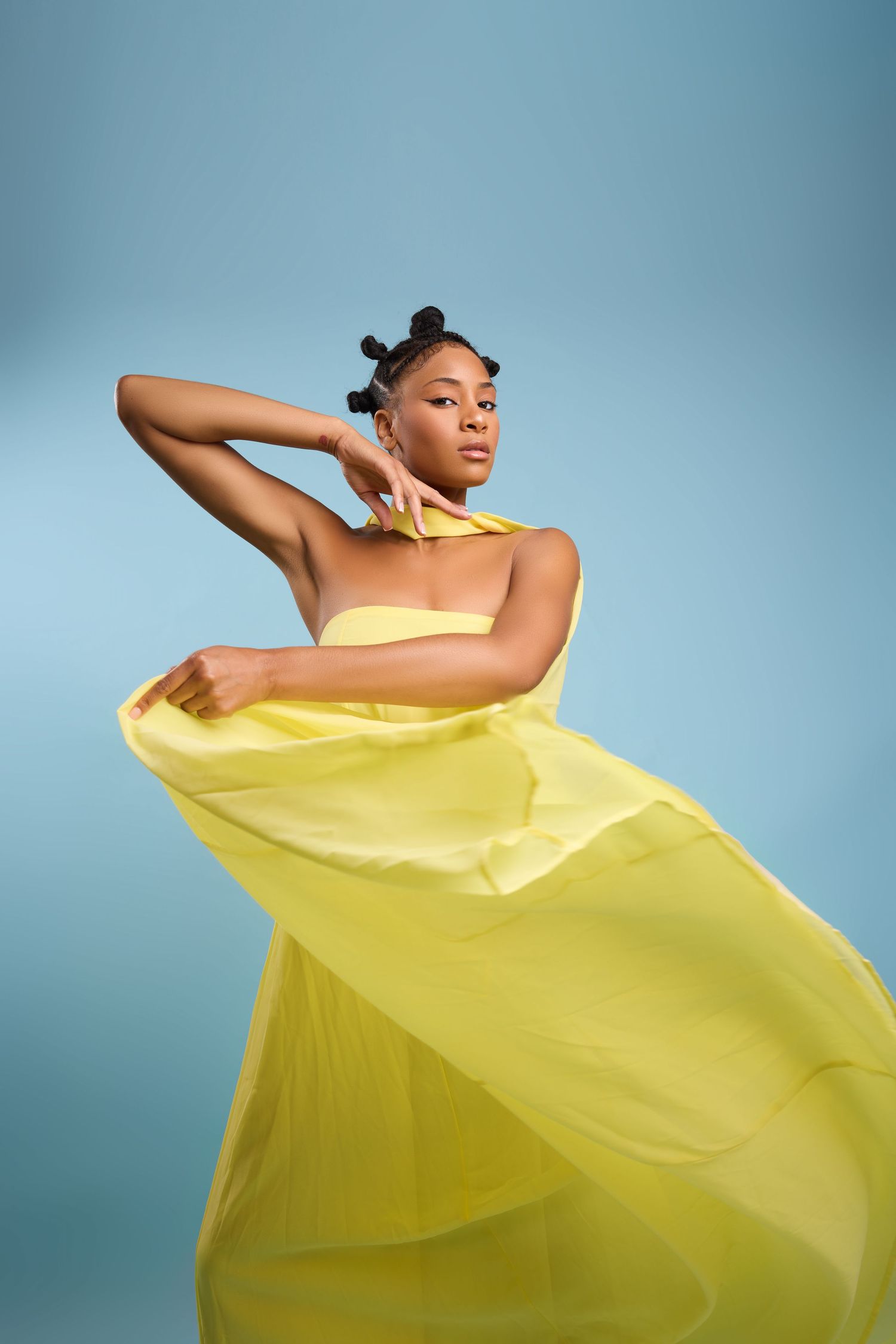 Figure in stunning yellow one-shoulder dress with gold accessories poses elegantly against brown backdrop.