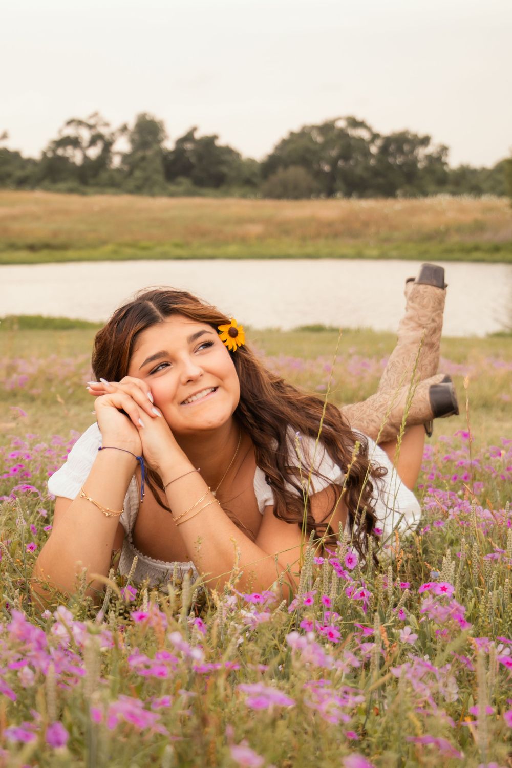 Girl laying in a flower field with a flower in her hair and a pond behind her.