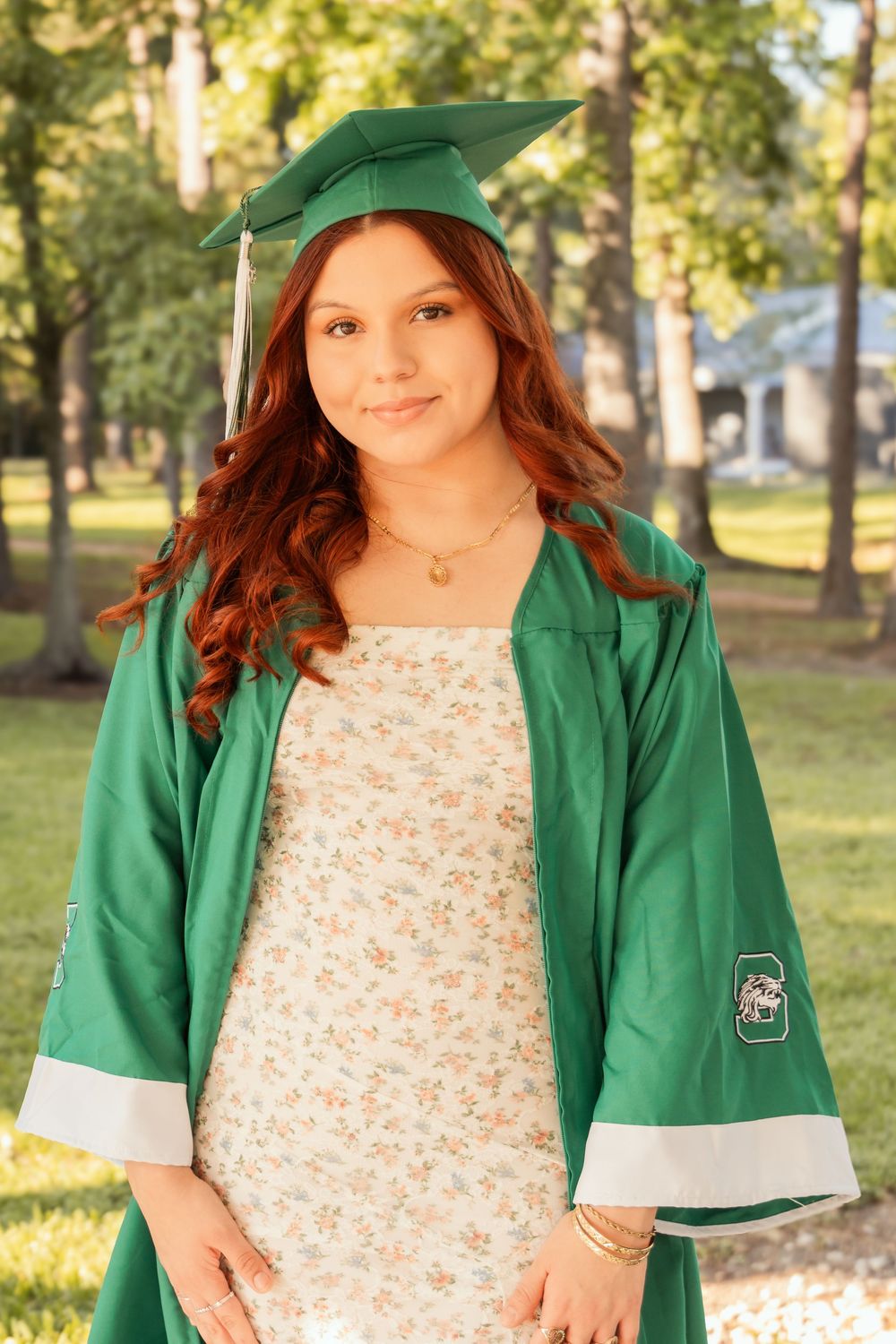 Photo of a high school senior in her cap and gown, smiling for graduation with a pretty outdoor background.