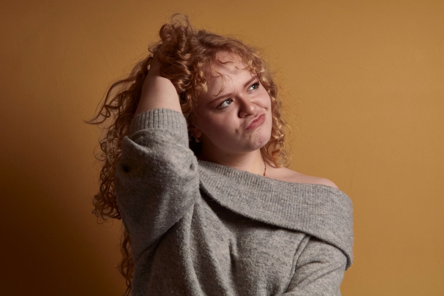 Studio portrait of a woman, mid shot of a goofy look and she has curly hair that she is holding up with one hand.