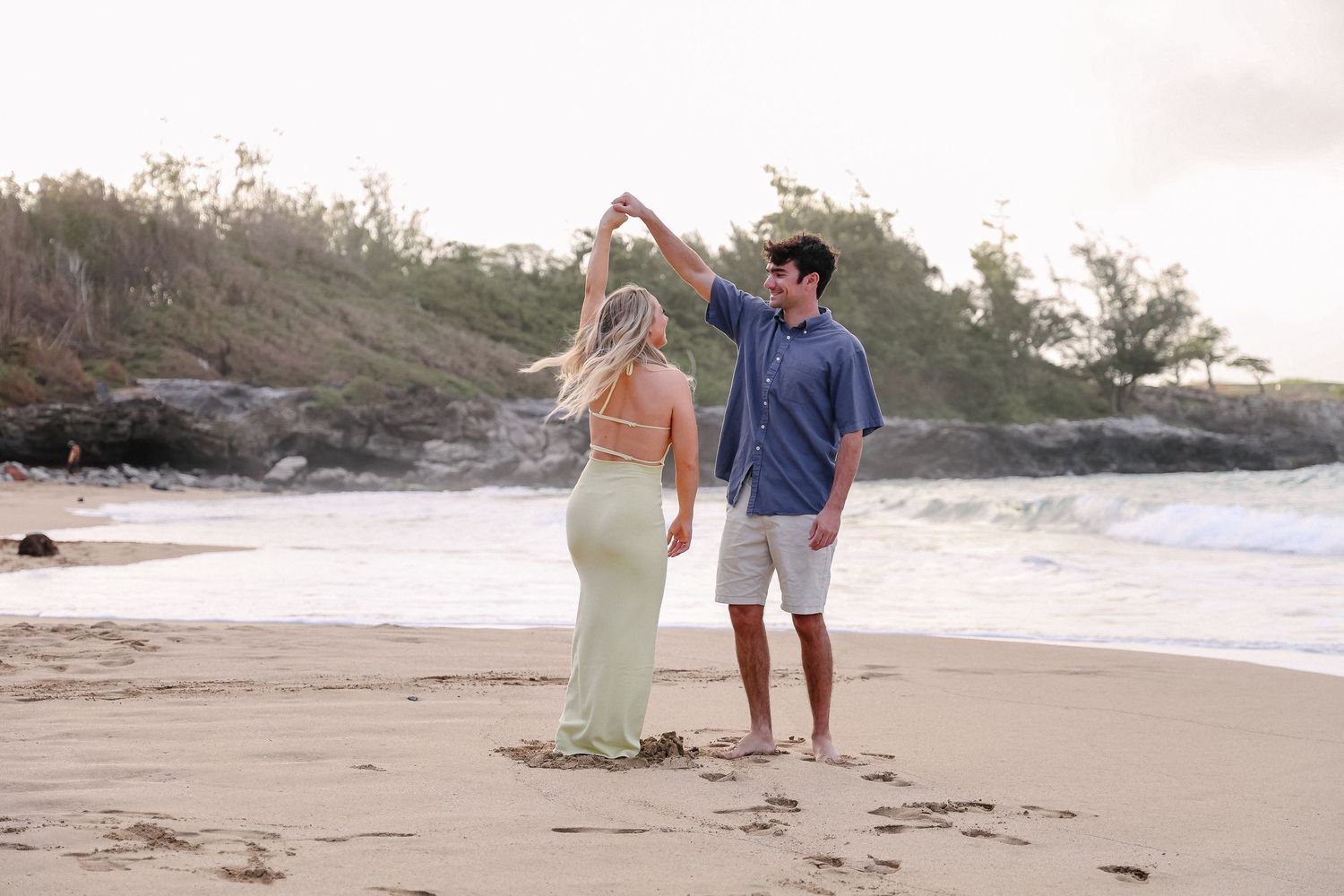 A couple walks along a sandy beach at sunset holding hands with ocean waves and rocky coastline in the background.