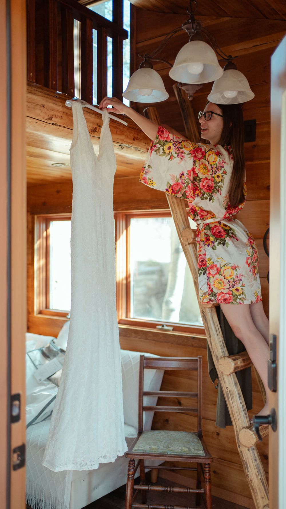 A white wedding dress hangs in a rustic wooden cabin while a bride in a floral robe stands on a ladder.