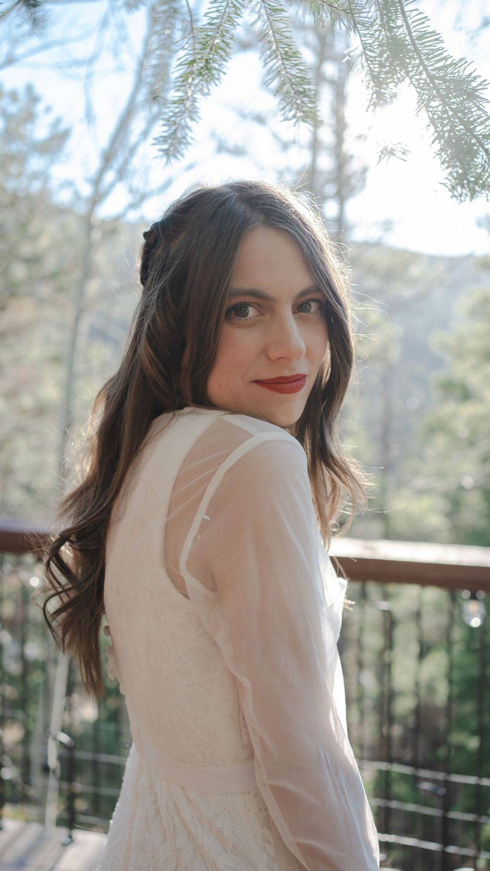A bride in a white dress poses on a deck surrounded by pine trees in soft natural lighting.