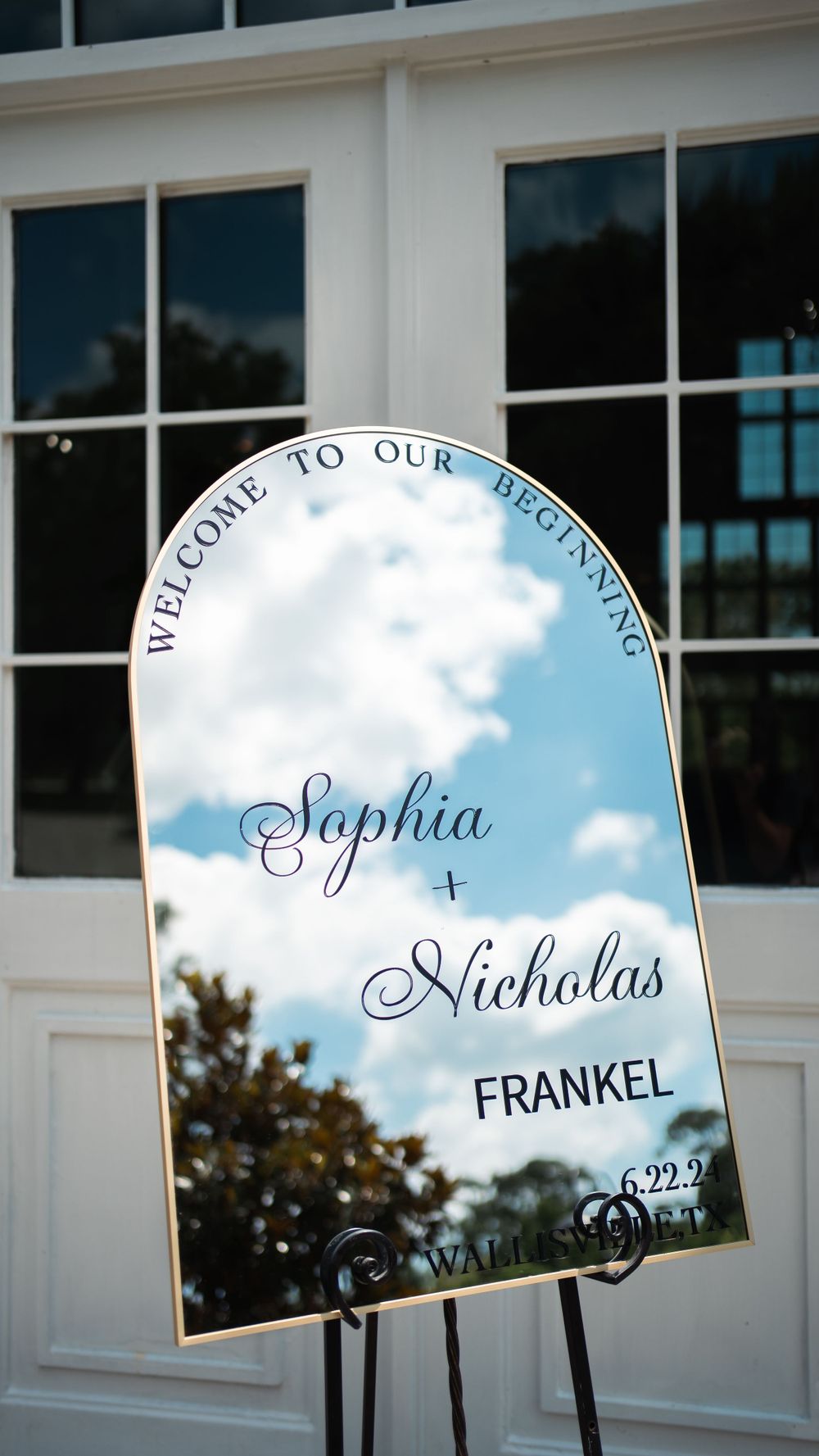 Mirrored wedding welcome sign with elegant calligraphy displaying Sophia and Nicholas Frankel's names against a cloudy blue sky.