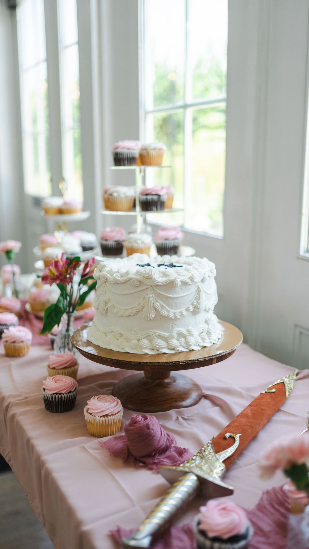 Elegant white wedding cake and pink cupcakes displayed on dessert table with floral decorations.