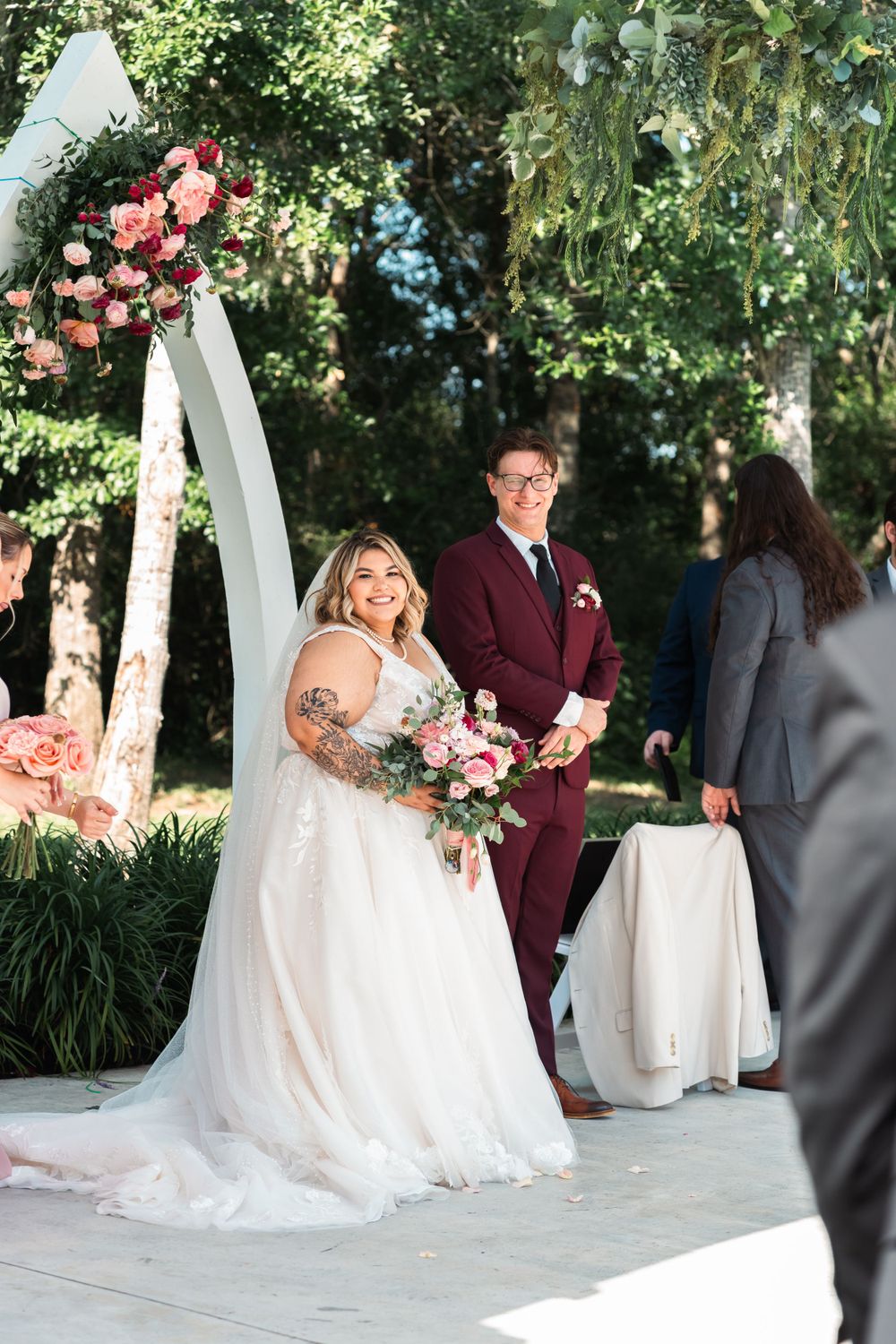 A wedding ceremony takes place under a white arch decorated with pink roses in a garden setting.