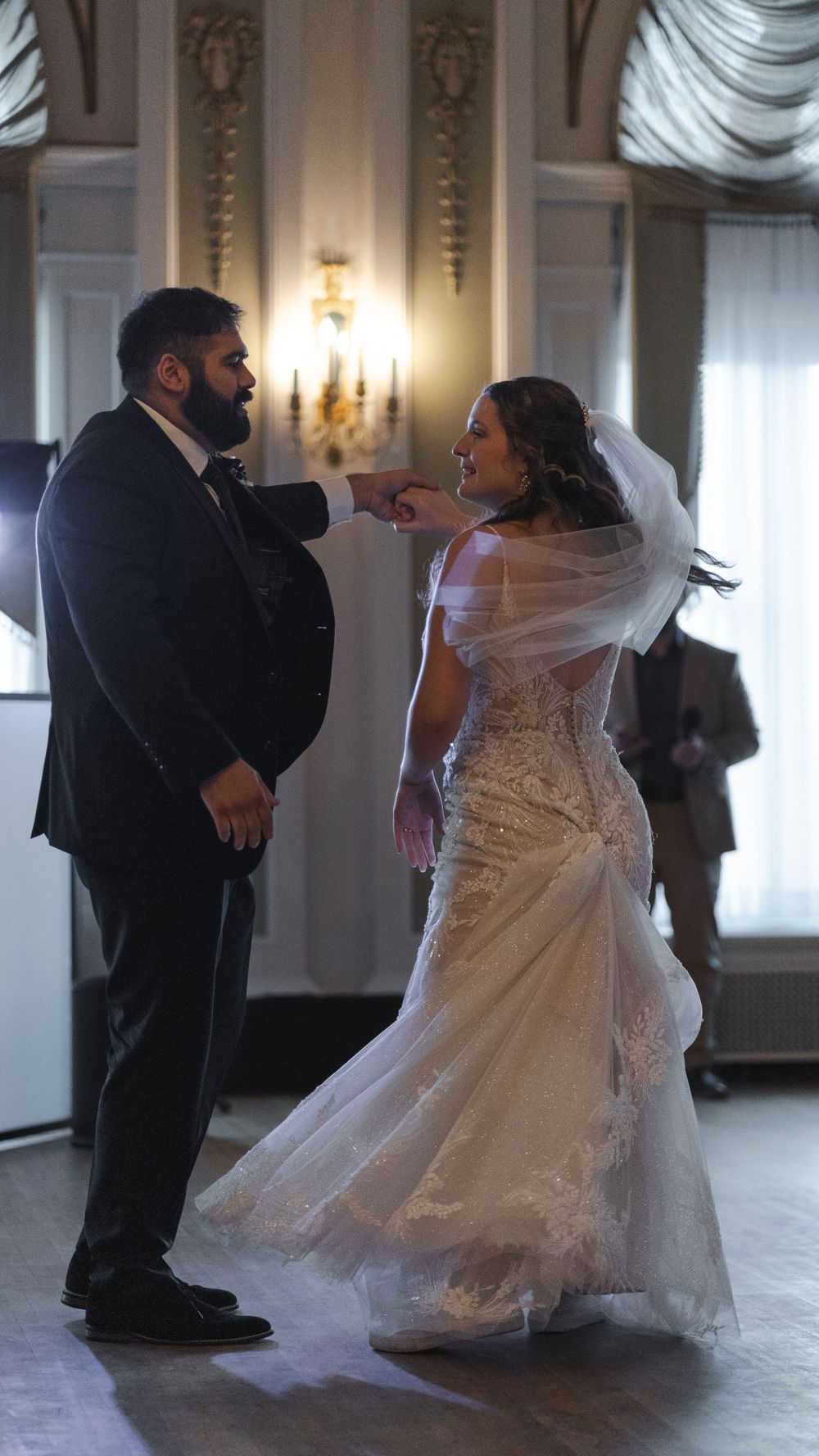A couple in formal wedding attire sharing a playful moment during their first dance in an elegant ballroom.