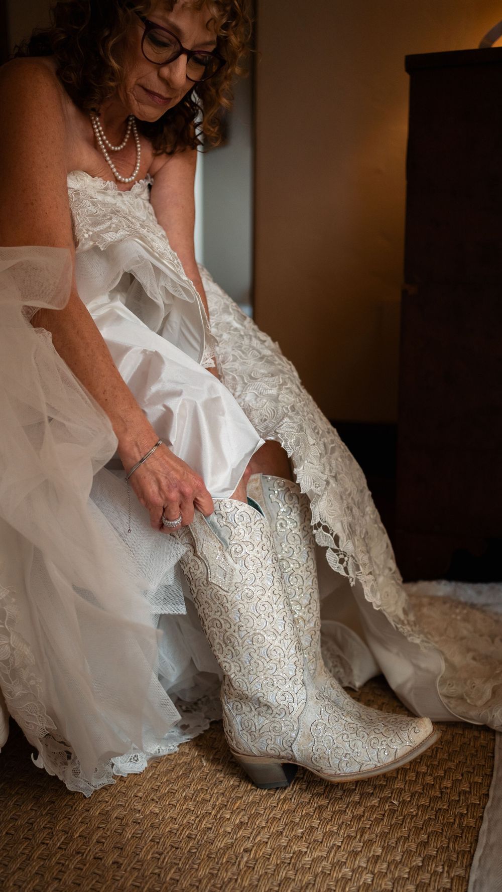 A bride adjusts her white lace cowboy boots while wearing a flowing lace wedding gown.
