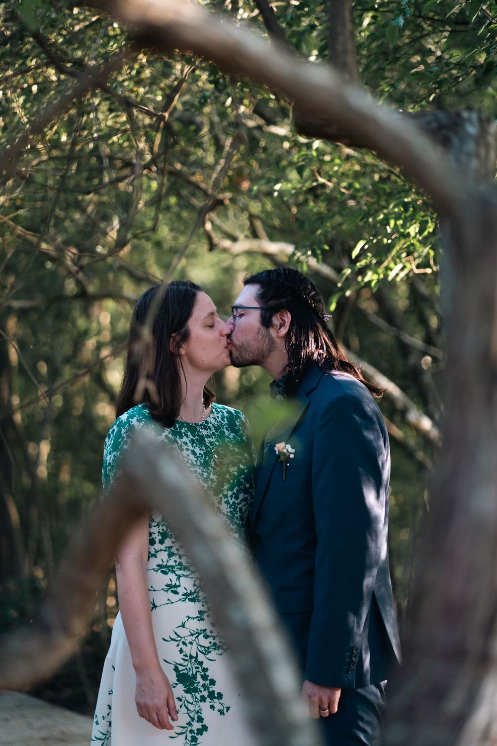 A couple shares a romantic kiss in a wooded outdoor setting with natural sunlight filtering through tree branches.