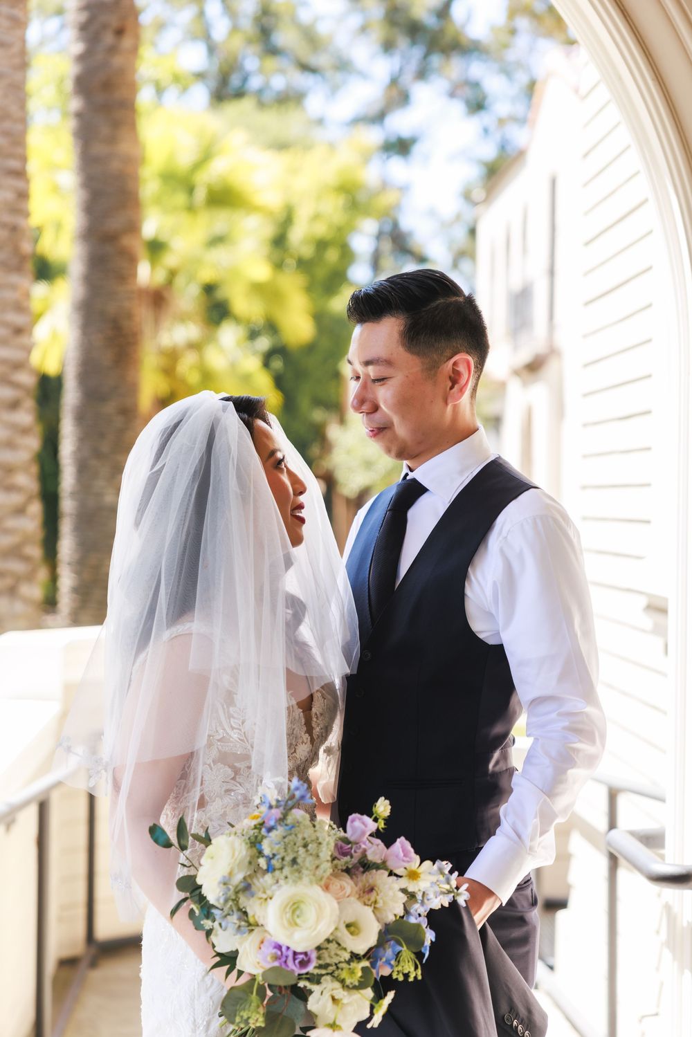 A wedding party stands outdoors near white columns, with the bride holding a bouquet of white and pastel flowers.