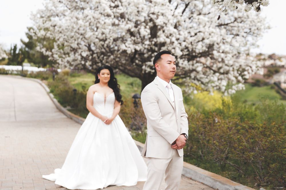 Couple in white wedding attire poses beneath blooming cherry blossom trees in a spring garden setting.