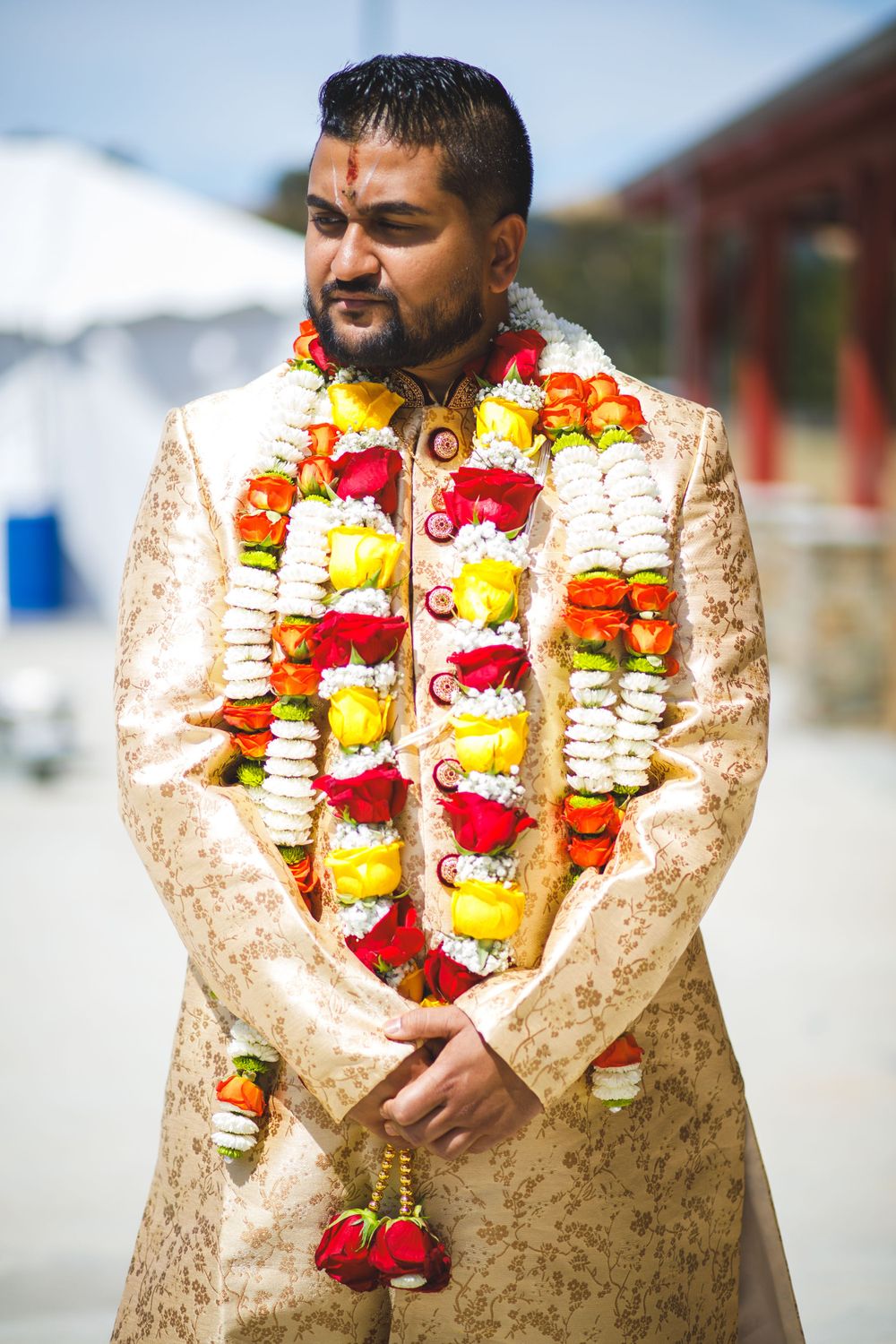 A groom wearing a gold embroidered sherwani adorned with colorful flower garlands at an Indian wedding ceremony.
