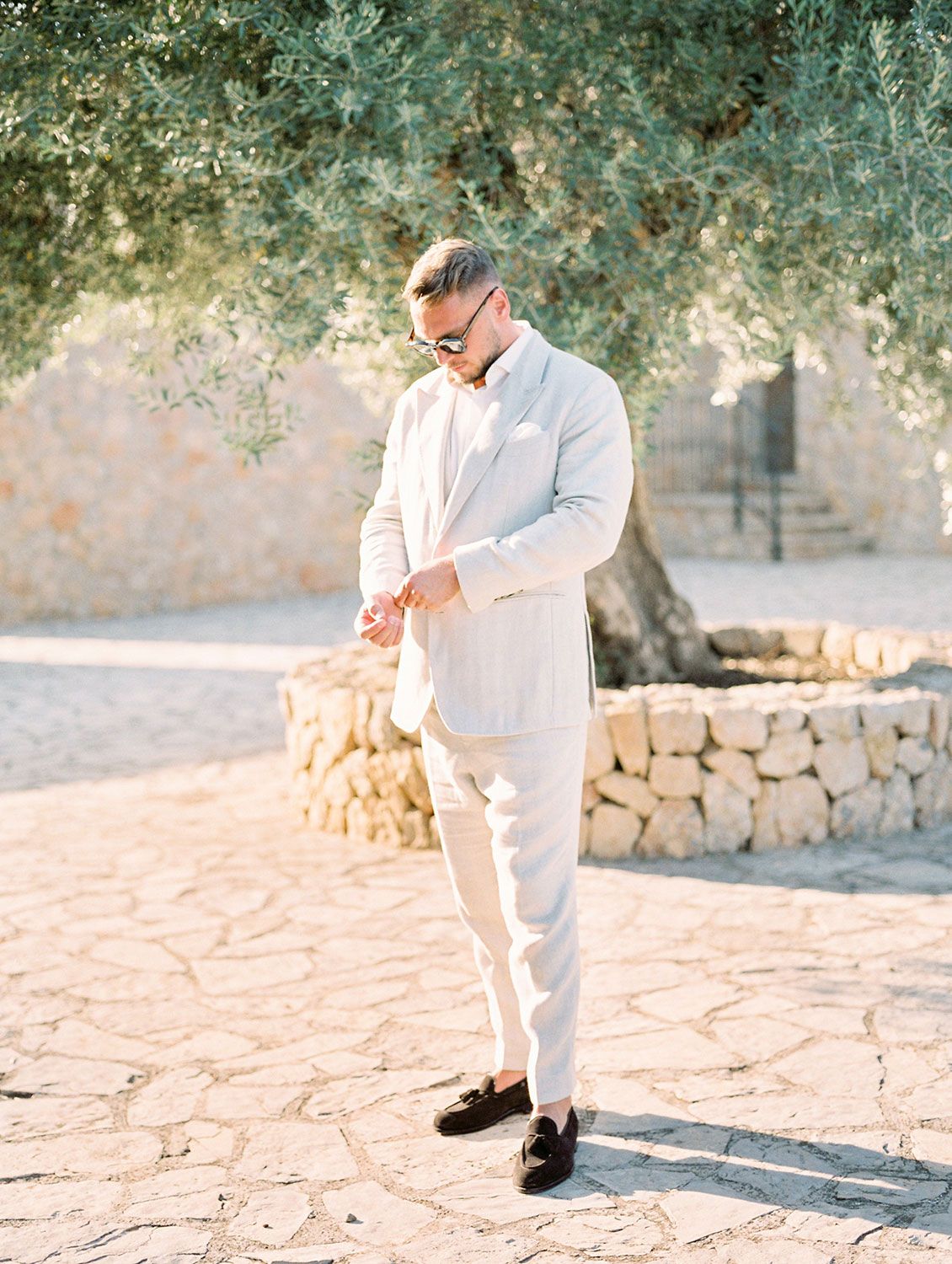 Stylish figure in an all-white outfit adjusts sleeve cuff while standing on stone pavers against Mediterranean backdrop.