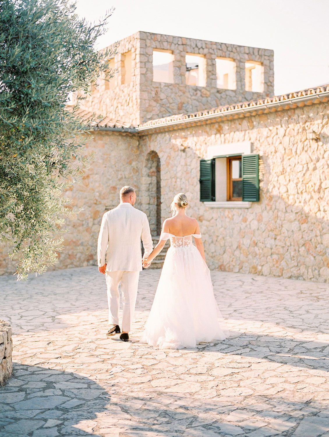 Light-filled stone villa with Mediterranean architecture serves as romantic backdrop for couple walking on cobblestone path.