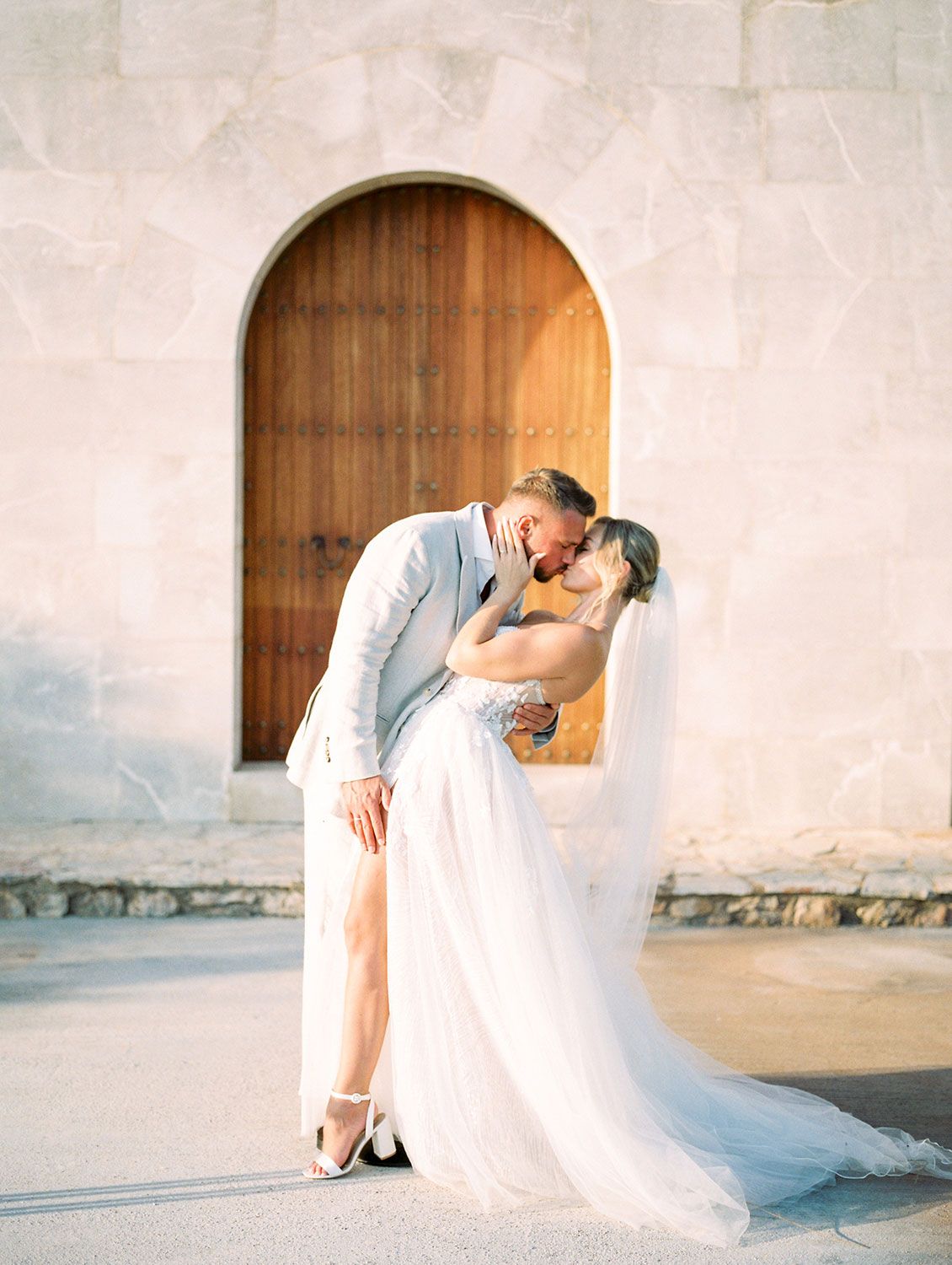 A romantic moment shared between a couple in wedding attire dipping in front of a rustic arched wooden doorway.