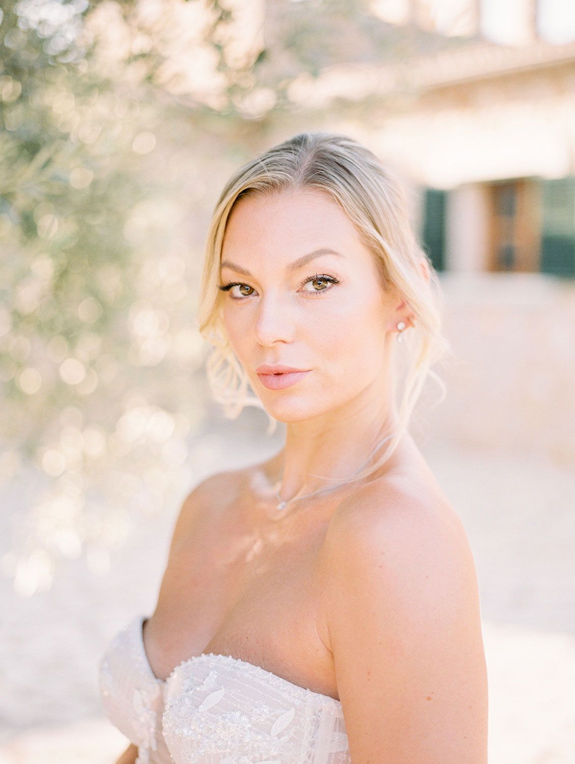 A bride poses outdoors in a strapless white beaded wedding gown with natural sunlight and soft bokeh background.