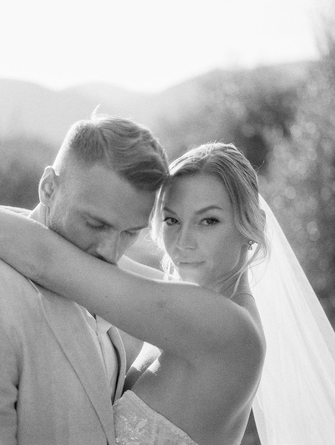 A romantic black and white wedding photo shows tender embrace between newlyweds in soft natural lighting outdoors.