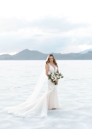 Bride in flowing white dress standing on salt flats with mountains in background.